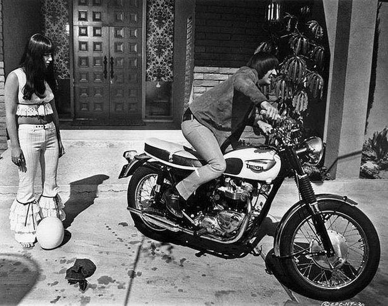 Sonny Bono mounts his Triumph Bonneville as Cher looks on.