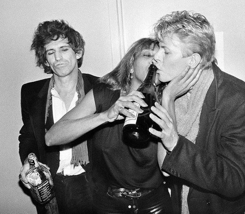 In 1983 at The Ritz New York, Tina Turner and David Bowie enjoy a champagne toast as Keith Richards observes with his whiskey bottle in hand.