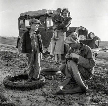 Family in California watches as man repairs flat tire on highway in 1937.