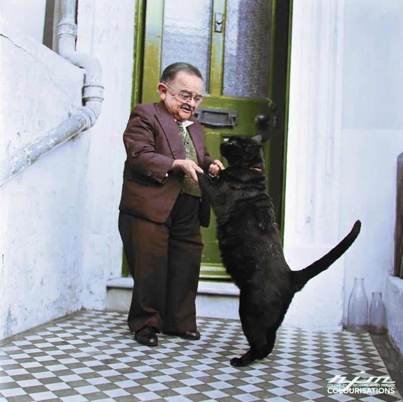 1956: Henry Behrens, the world's tiniest man, joyfully dances with his pet cat at the entrance of his Worthing residence.